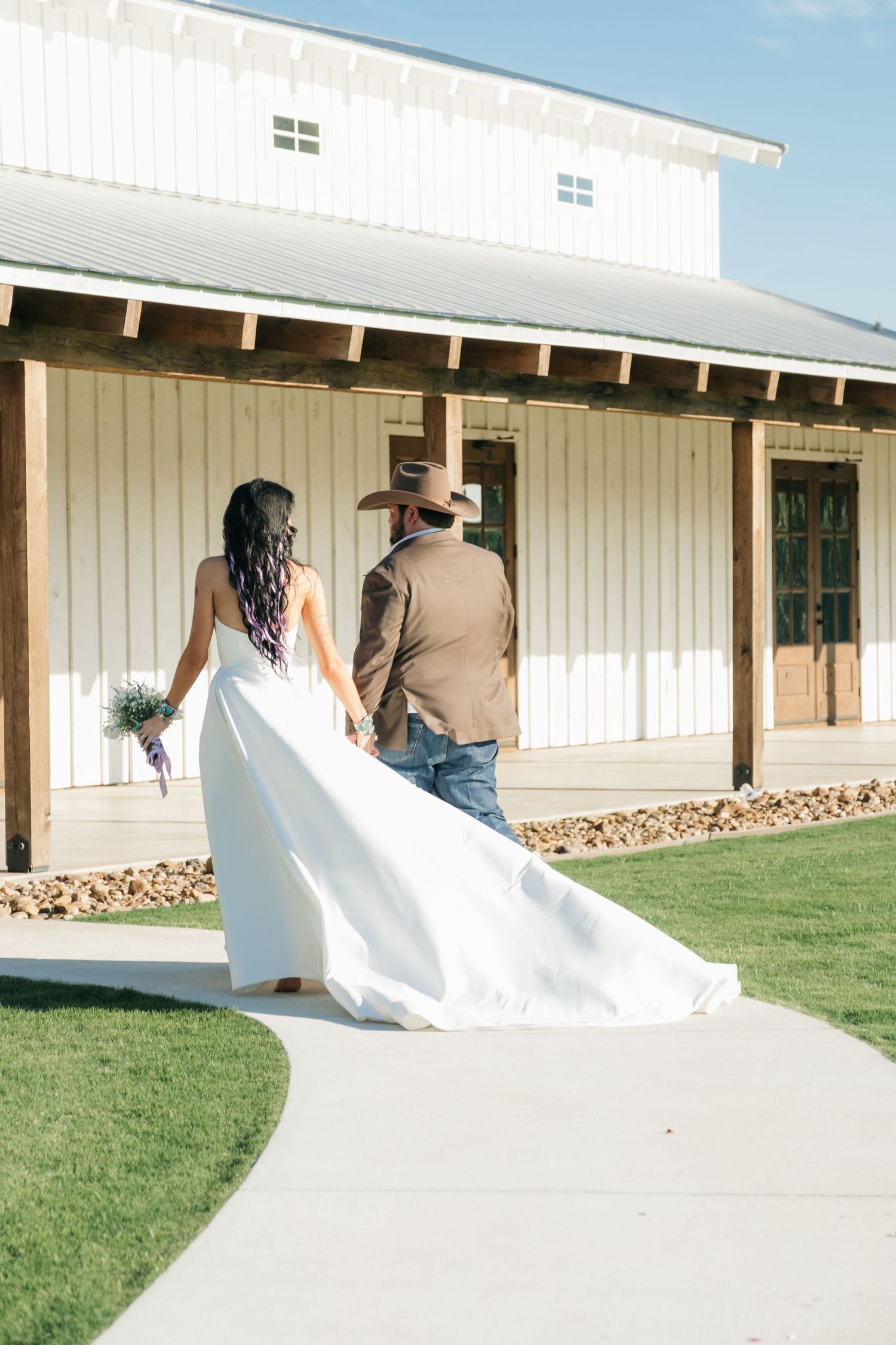 A couple walking on a path outside a white barn-style building with the brides flowing white dress trailing behind them.