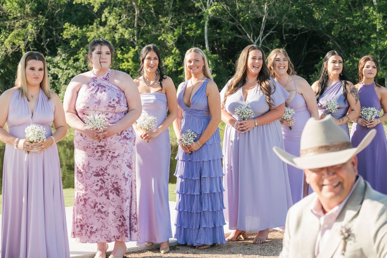 Wedding party dressed in coordinating lavender and floral dresses poses together in an outdoor garden setting.