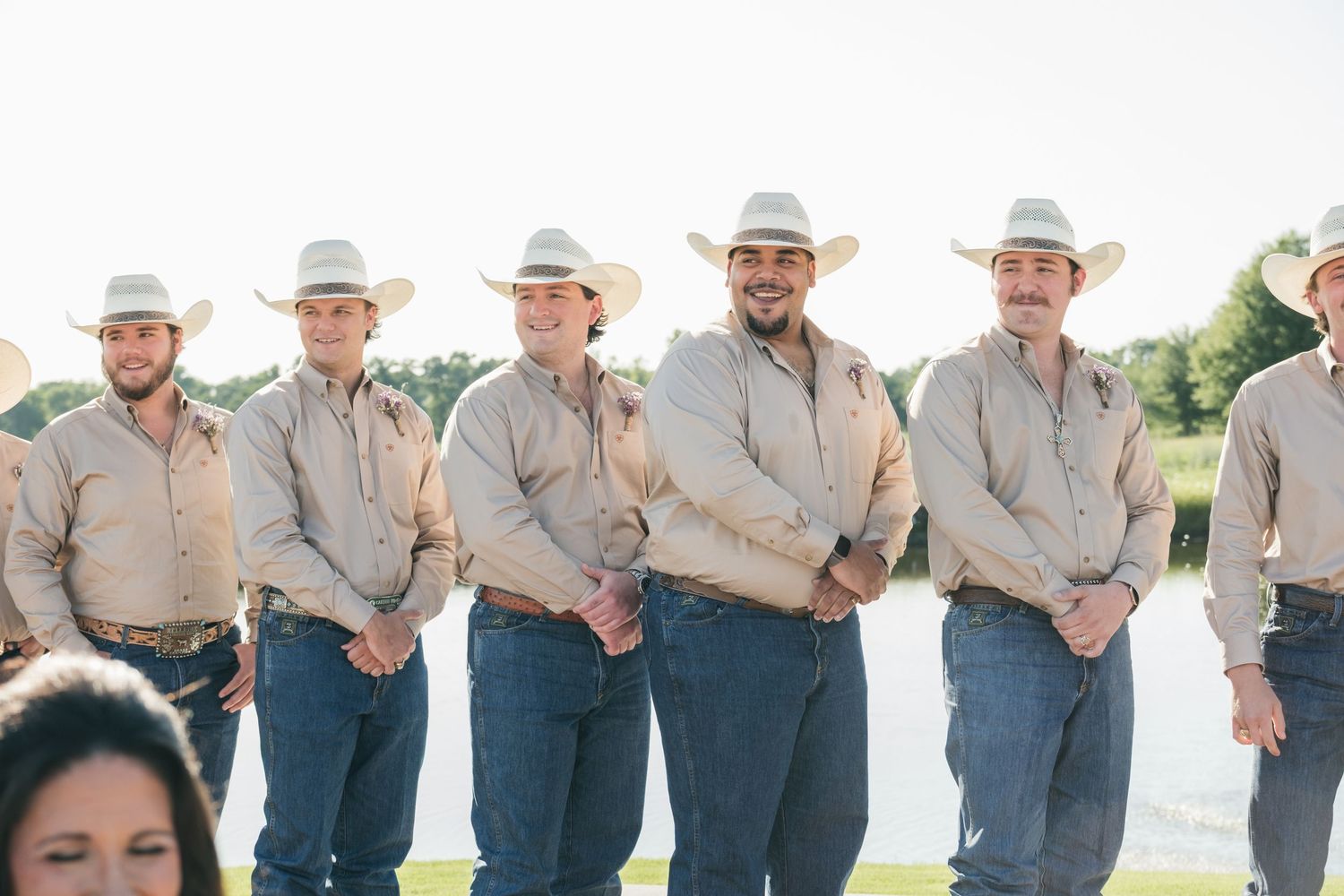 Groomsmen wearing matching light colored shirts and cowboy hats stand in a line by a lake.