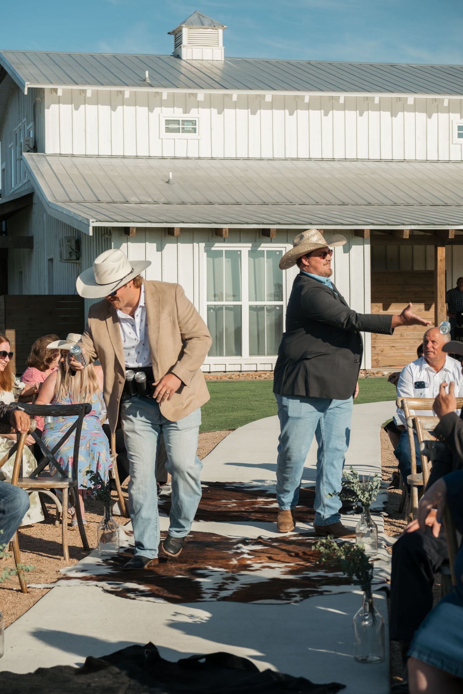 Two men in cowboy hats stand on a stage in front of a white building during an outdoor gathering.