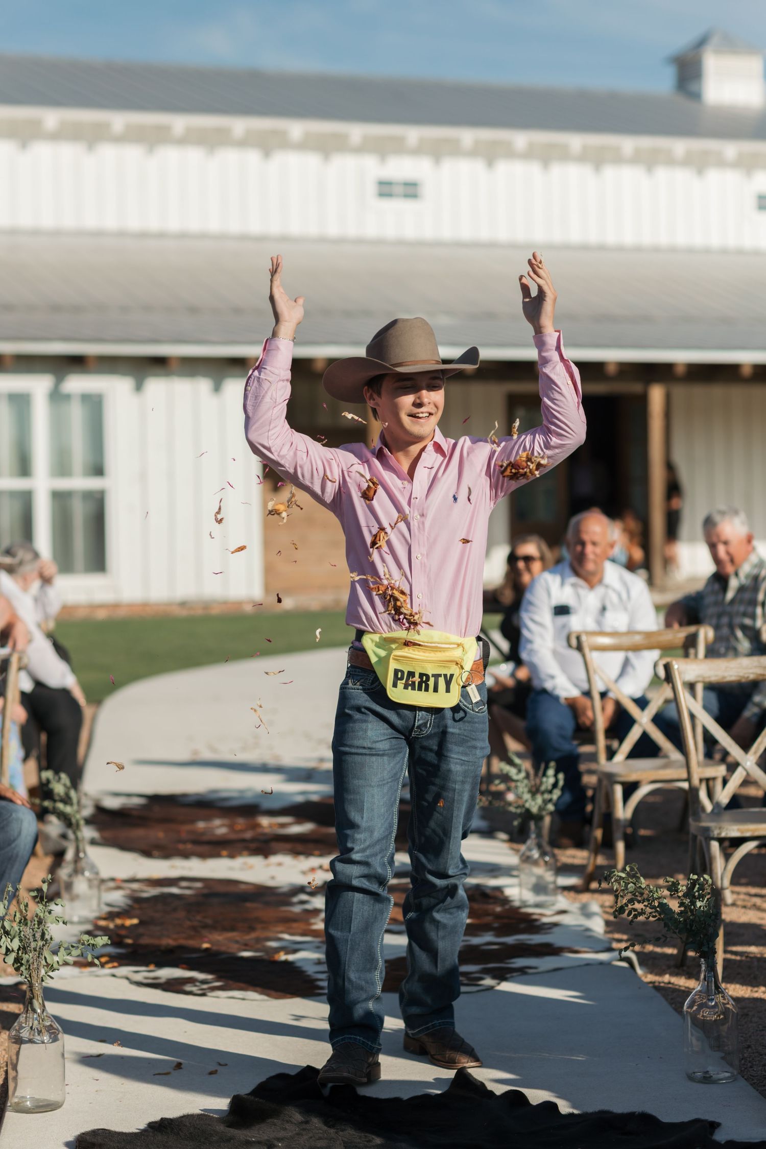 Person wearing pink button-down shirt and jeans joyfully dances outdoors at a sunny daytime event with tables in background.