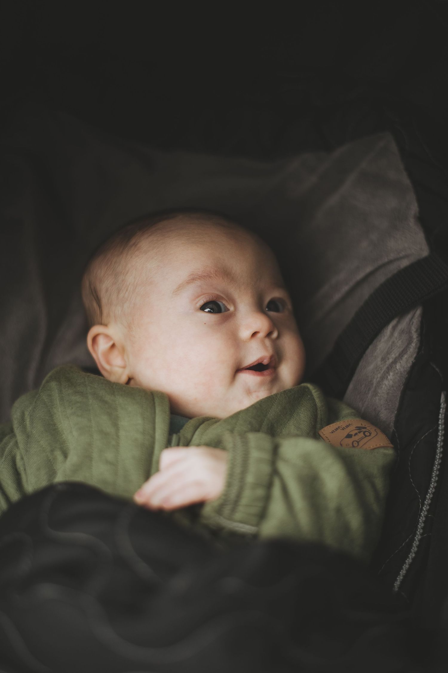 Baby wearing green sweater looks up with bright eyes while nestled in dark car seat or stroller.