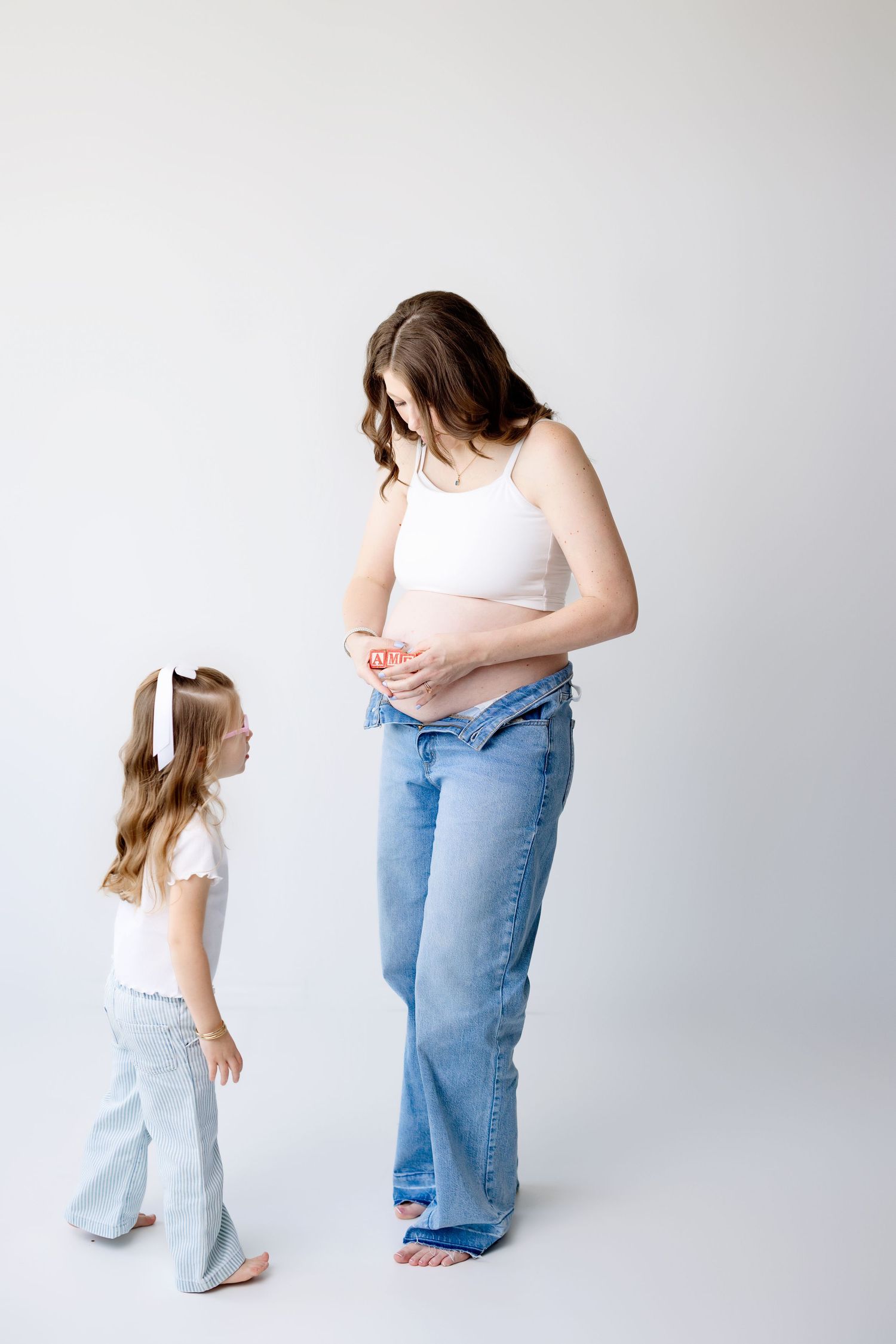 Against a white backdrop, a woman and young child stand facing each other in matching light denim jeans and casual attire.