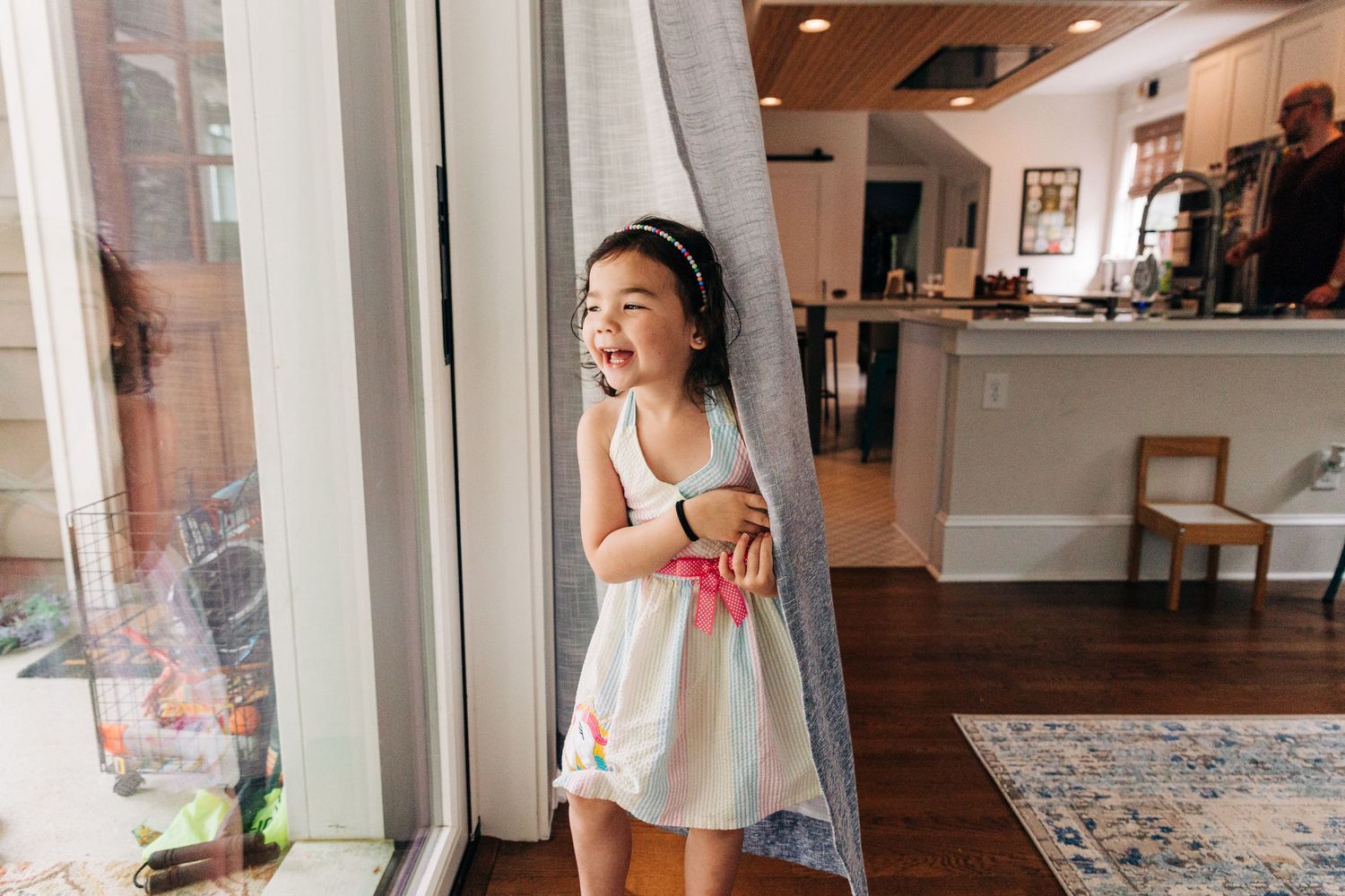 Young girl in white dress holds apple by window in sunny living room with hardwood floors and colorful rug.
