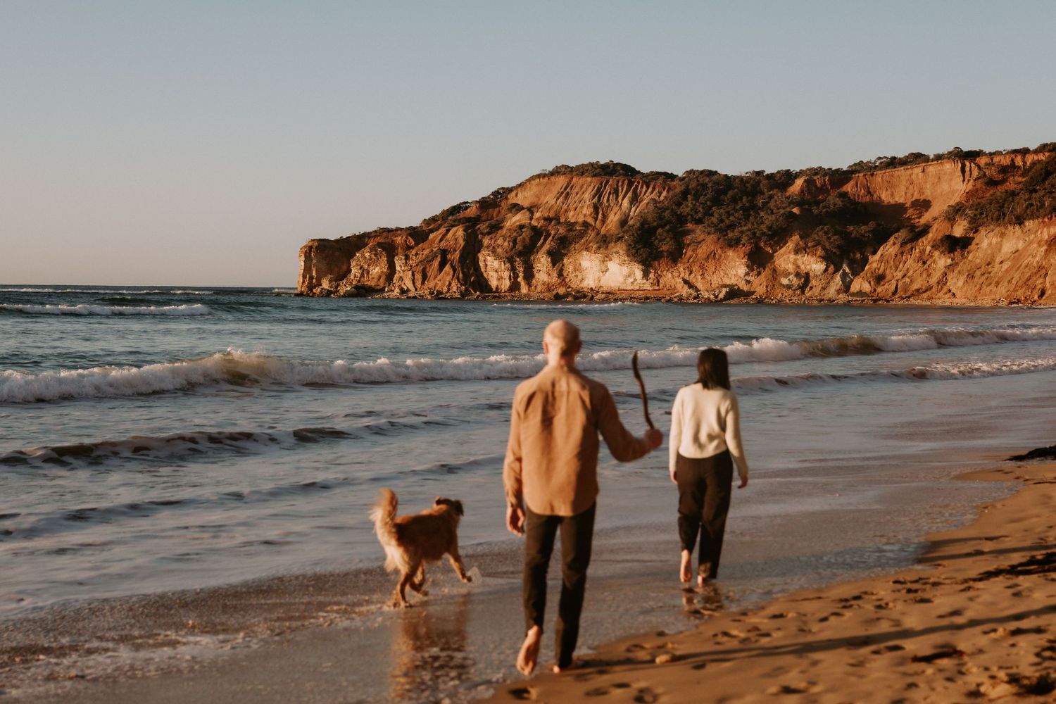 People walking with a dog on a sandy beach at sunset with dramatic coastal cliffs in the background.