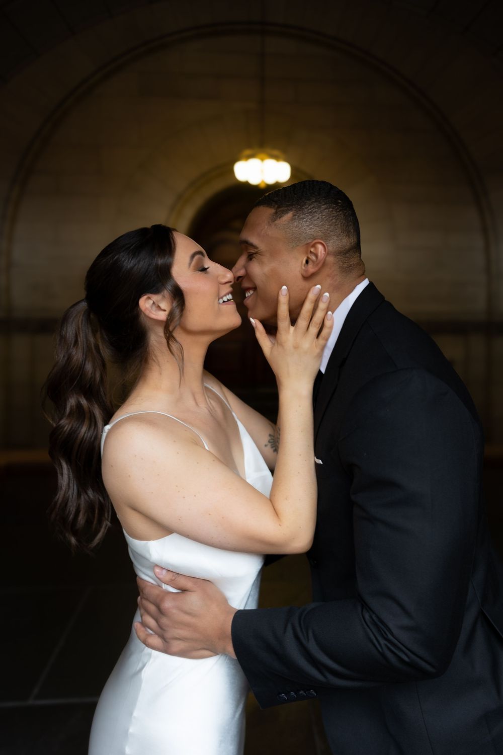 A romantic wedding couple sharing an intimate moment under an arched hallway with soft lighting.