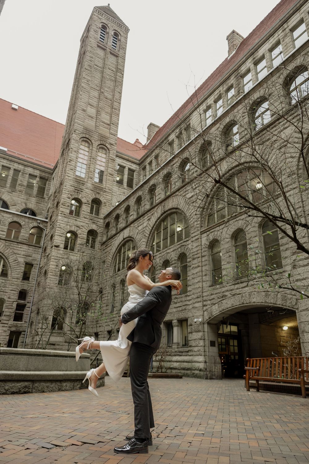 A couple celebrates their engagement in front of a grand stone building with Gothic architecture and a towering clock tower.