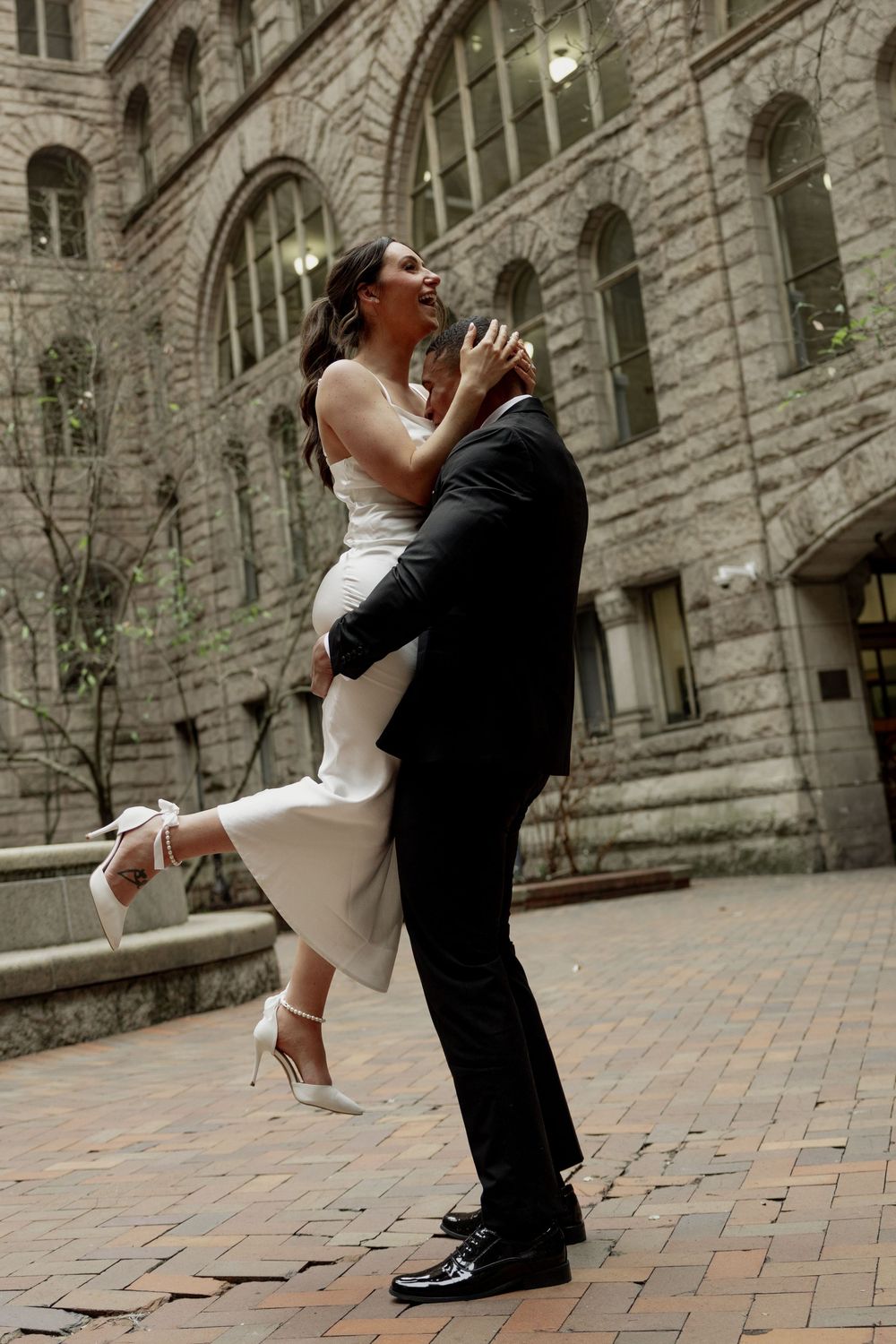 A couple shares a romantic embrace on a brick courtyard in front of a historic stone building with arched windows.
