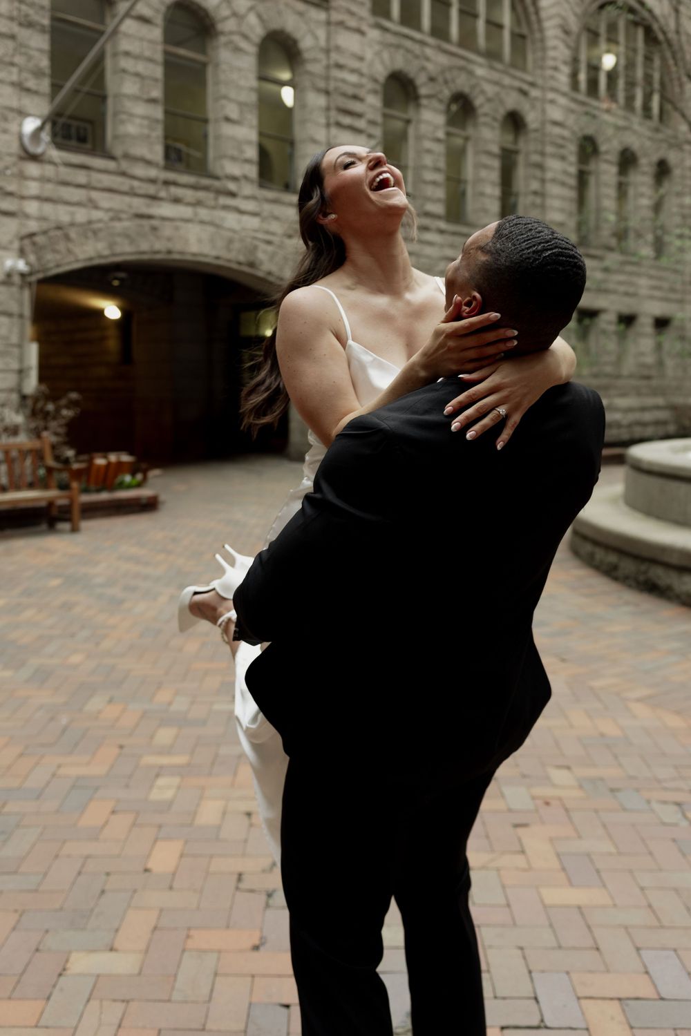 Joyful newlyweds embrace and laugh together on a brick courtyard in front of a historic stone building.