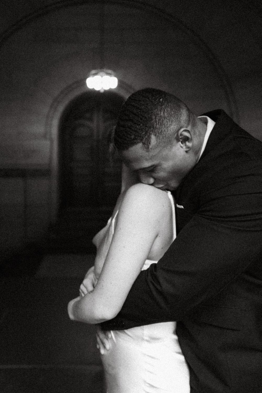 A black and white intimate photograph showing a tender embrace in dramatic lighting with arched architecture visible.