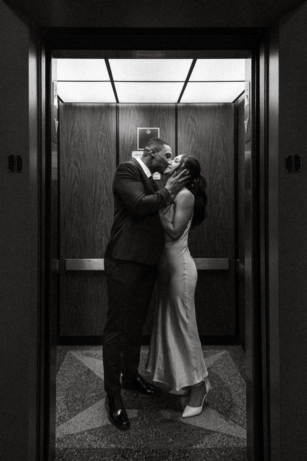 A romantic black and white moment captured in an elevator as a couple in formal attire share an intimate kiss.
