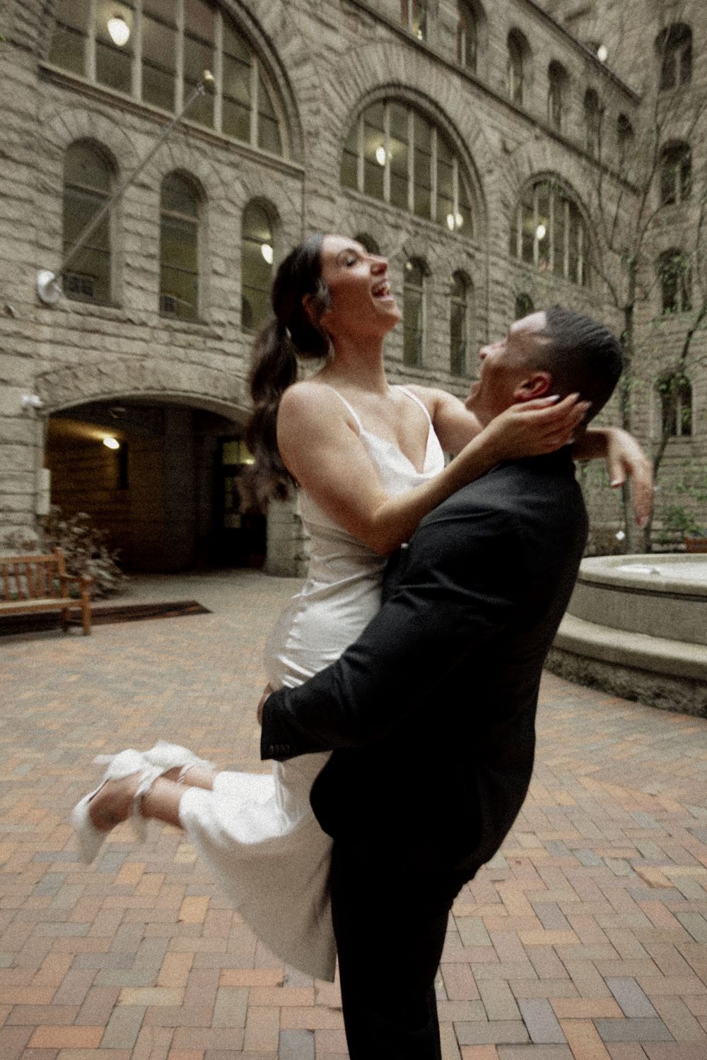 A romantic moment between newlyweds embracing in front of a historic stone building with arched windows and brick courtyard.