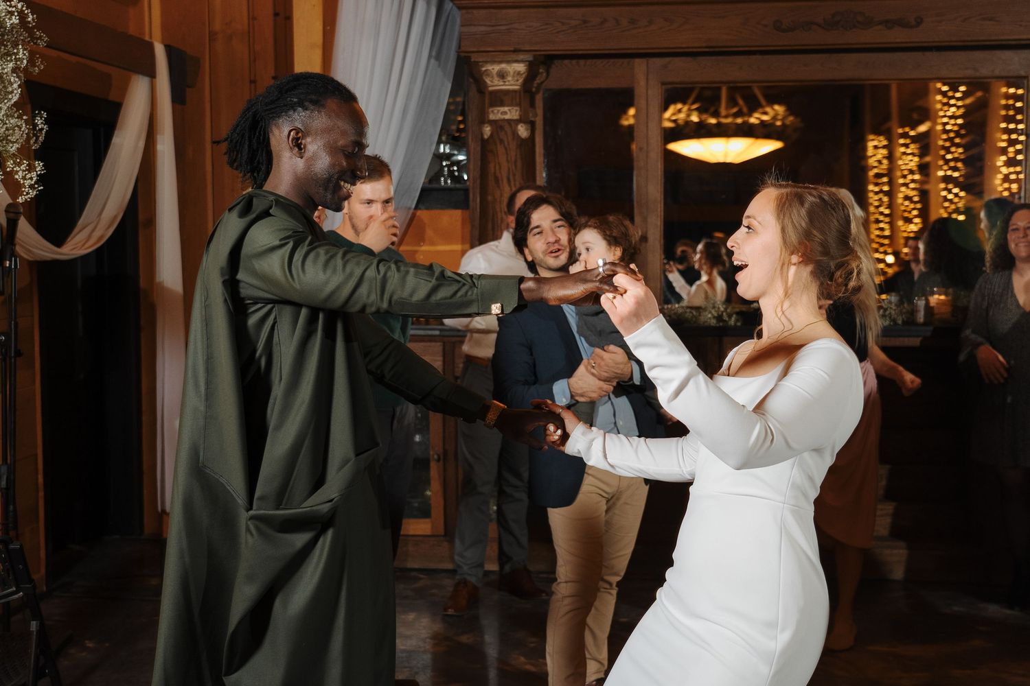 People dance energetically at a reception venue with indoor lighting and decorative elements in the background.