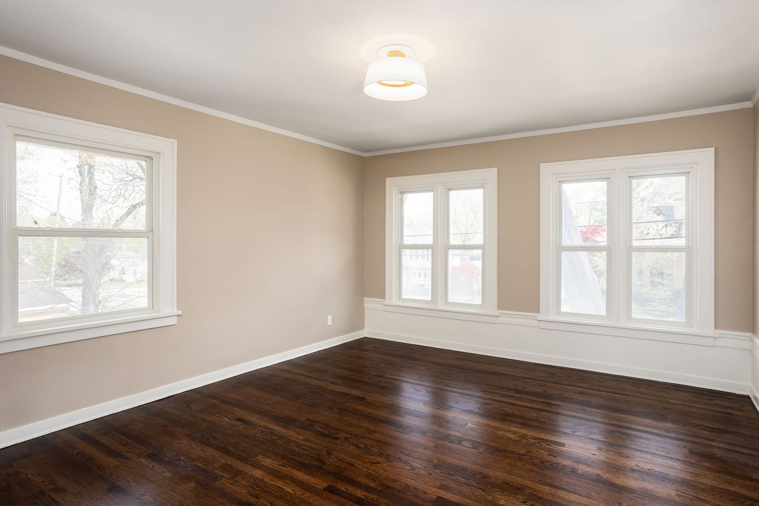 Empty room with dark hardwood floors, beige walls, and white trim featuring multiple windows and ceiling light fixture.