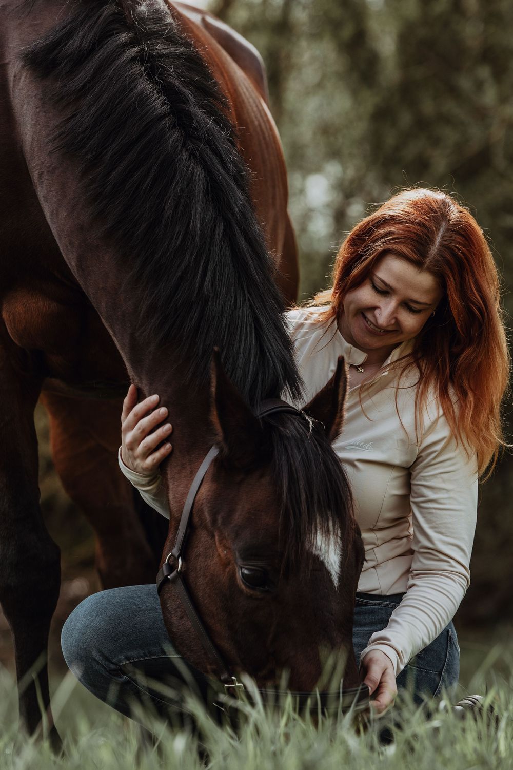 A person in a white sweater gently pets a dark brown horse while sitting in tall grass at dusk.