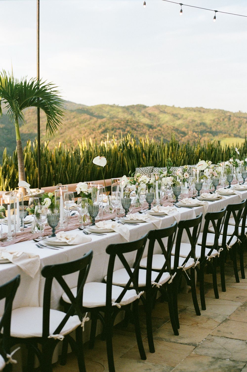 Long wedding reception table overlooking mountain landscape with string lights overhead.
