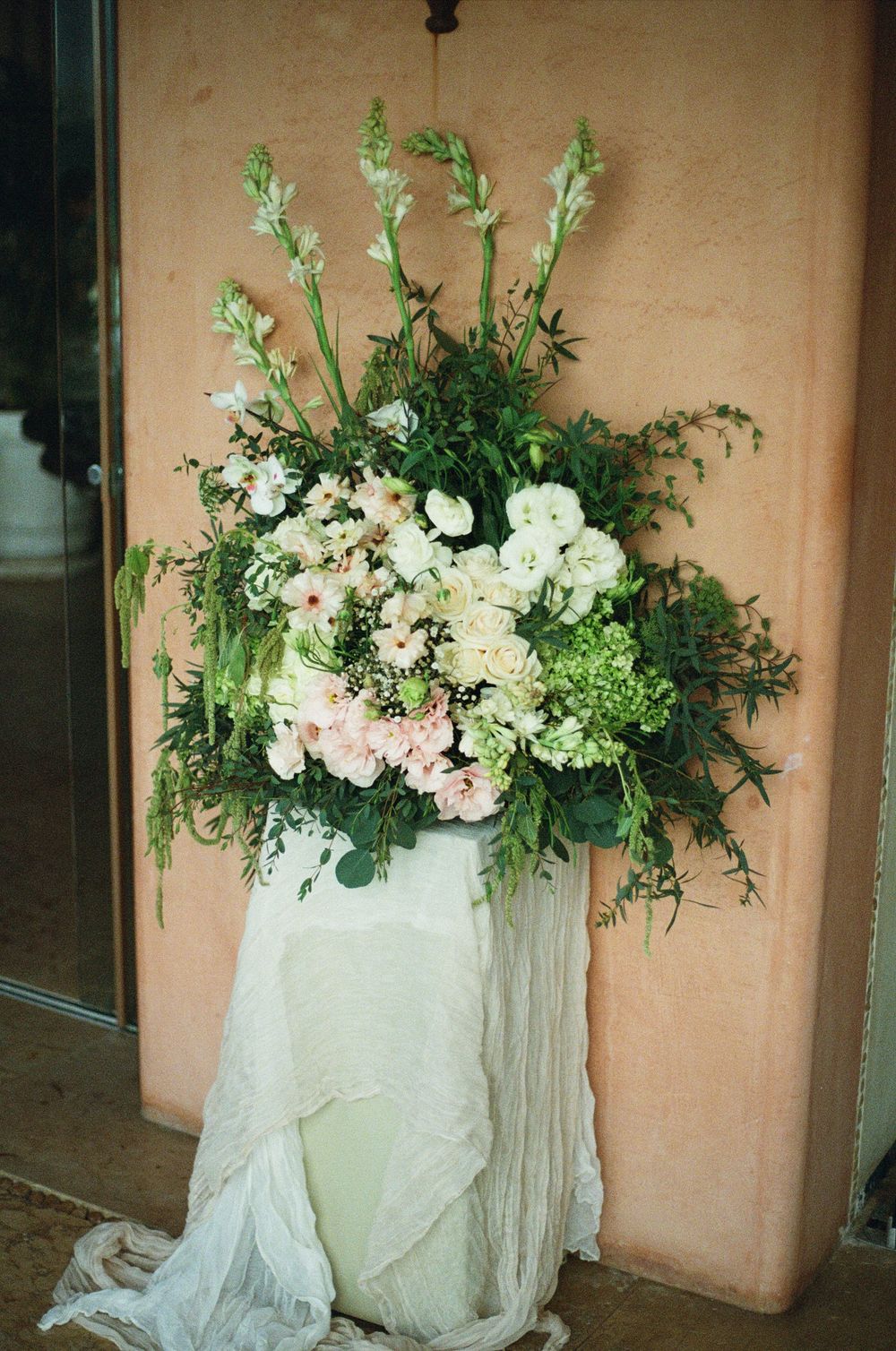Elegant white floral arrangement with greenery displayed on a draped pedestal against a peach wall.