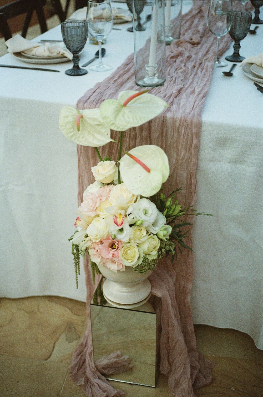 Wedding table centerpiece featuring white roses and blush fabric runner on mirrored stand.