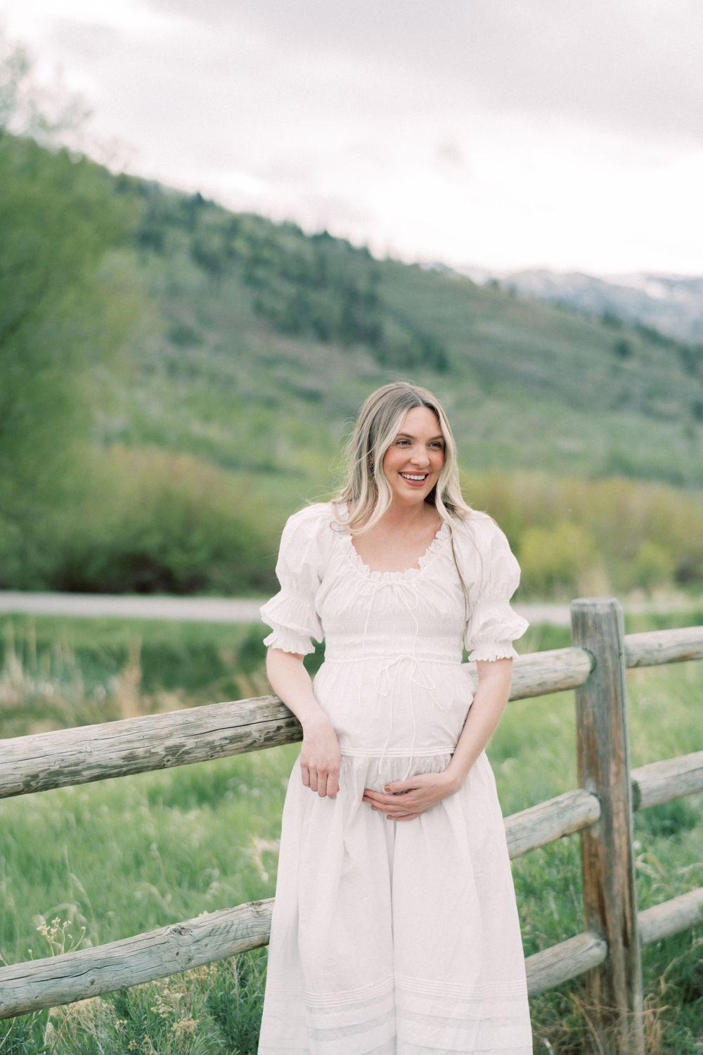 Pregnant woman in flowing white dress poses by rustic wooden fence with mountain backdrop during springtime.