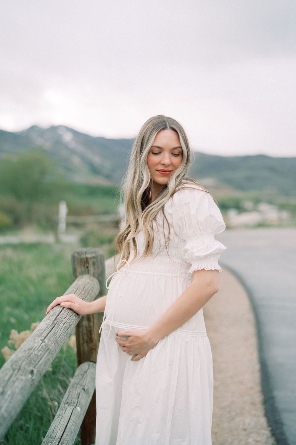 A person in a flowing white dress stands by a rustic wooden fence with mountains in the background.