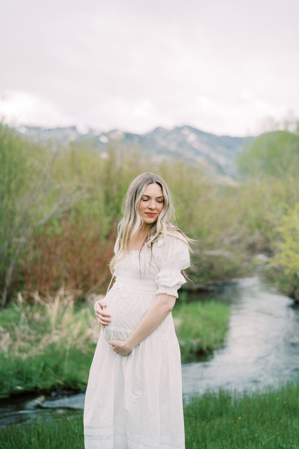 Expectant mother in white dress stands beside mountain stream during maternity photoshoot in scenic landscape.