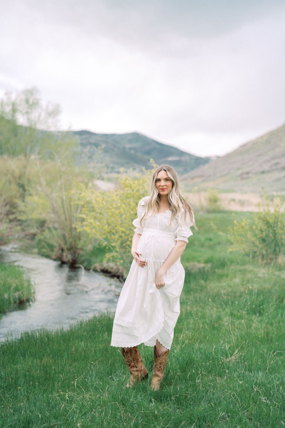 A person in a flowing white dress and brown boots stands in a green field by a stream with mountains in the background.