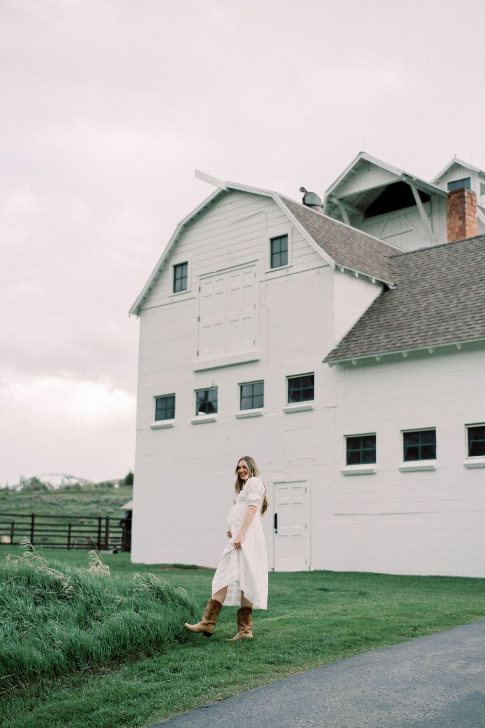 A figure in a flowing white dress stands outside a classic white barn in a pastoral rural setting against a moody sky.