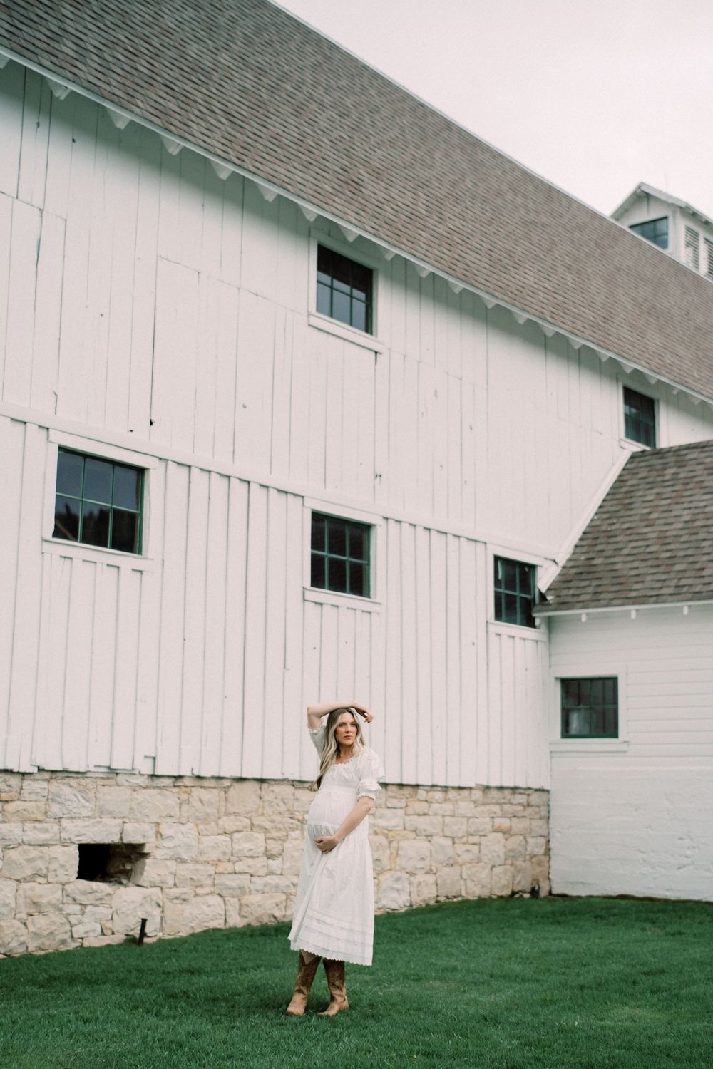 A pregnant woman in a white dress poses in front of a rustic white barn with stone foundation on a grassy lawn.