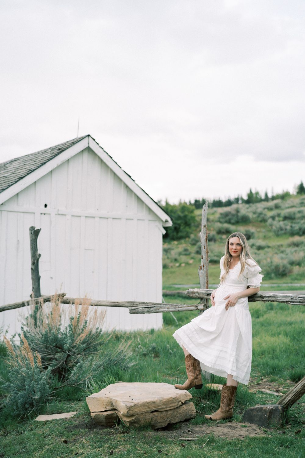 Rustic white farmhouse setting with figure in flowing white dress and boots standing by wooden fence in rural landscape.