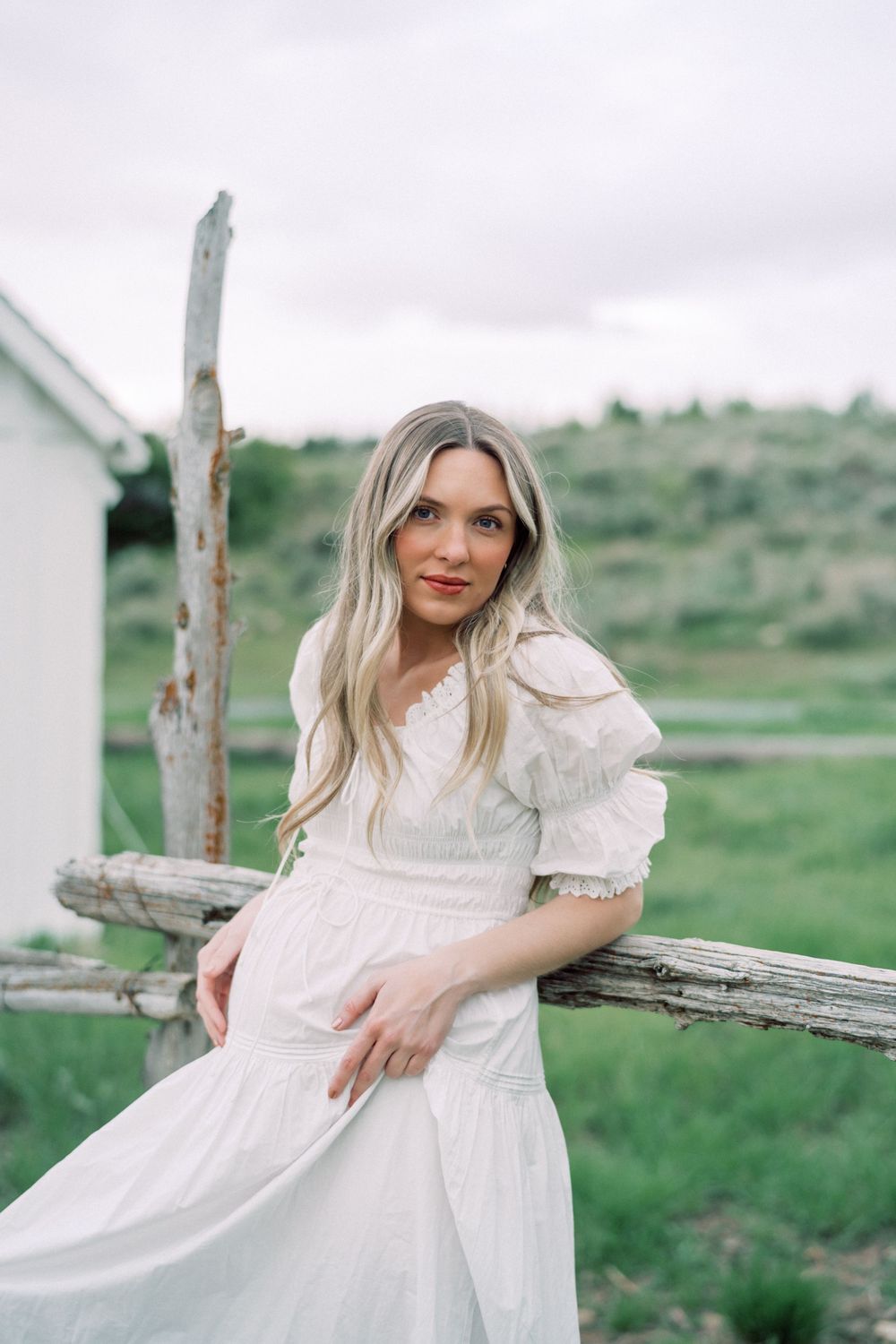 A person in a romantic white prairie dress stands against a rustic wooden fence in a pastoral meadow setting.