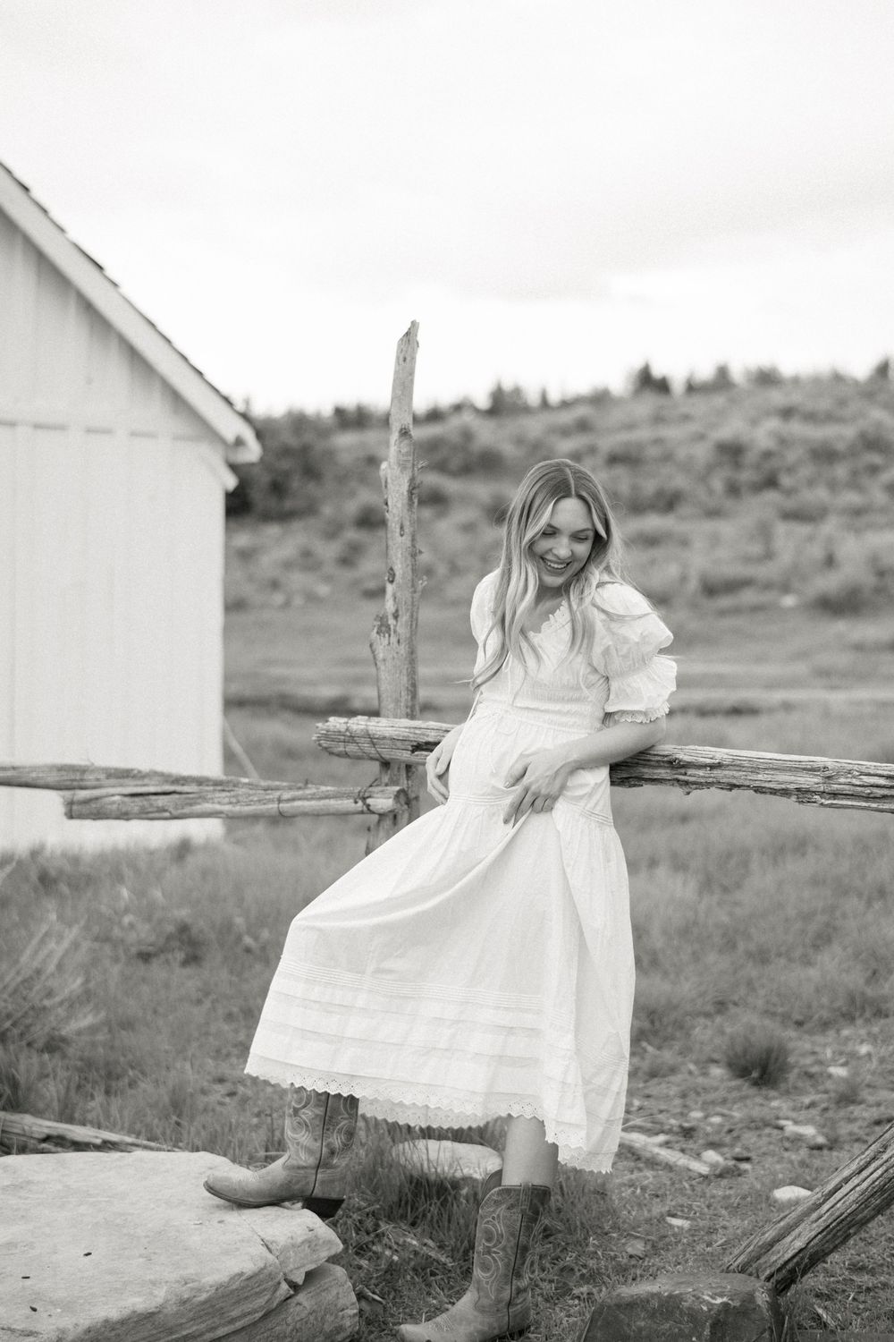 Black and white country photo showing someone in a flowing white dress and boots sitting on a rustic wooden fence near a barn.