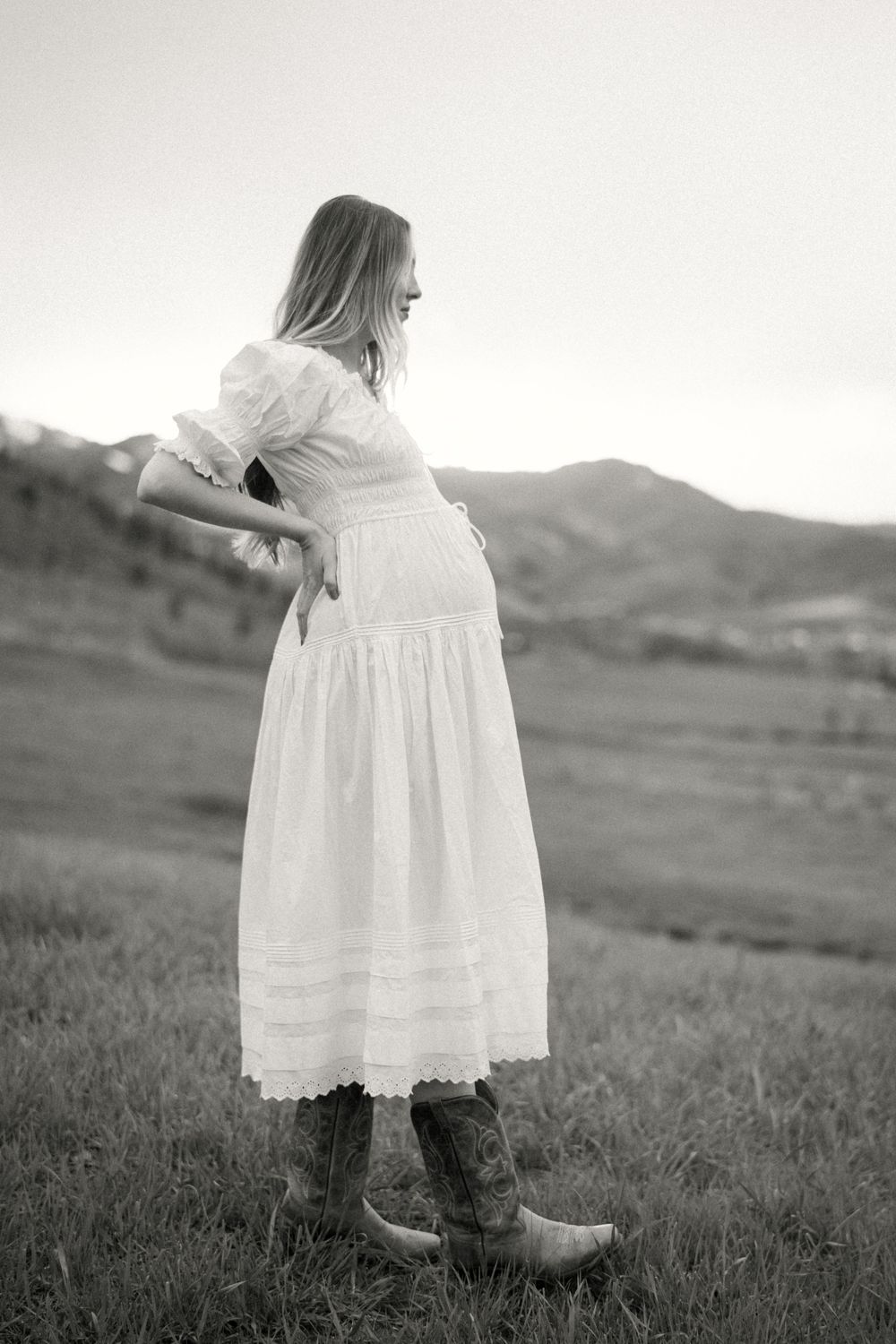 Black and white photo of someone in a flowing white vintage dress and cowboy boots standing in a grassy field.