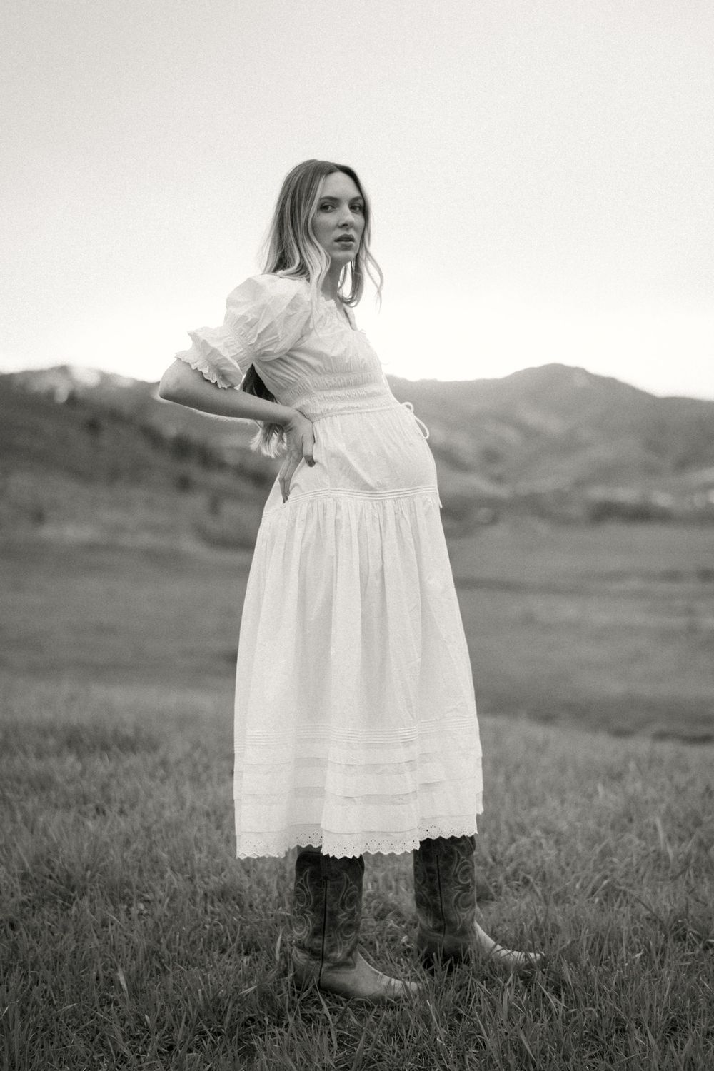 Black and white portrait of someone wearing a Victorian-style white dress and boots standing in a grassy field with hills.