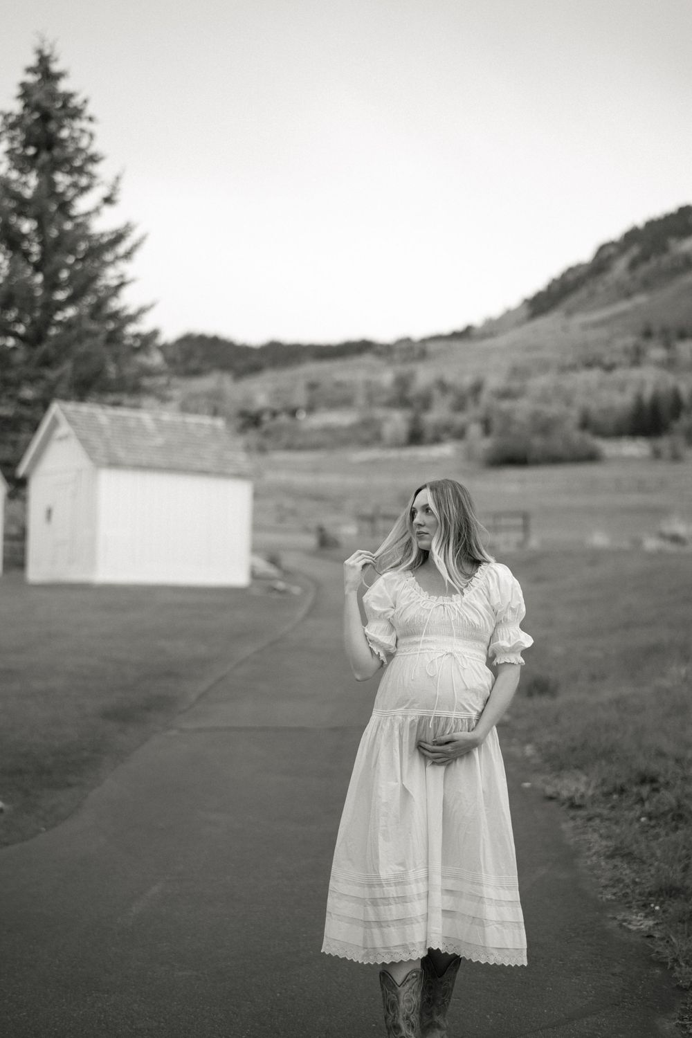 A vintage-style black and white photograph showing an expectant woman in a flowing white dress standing outdoors near a rustic shed.
