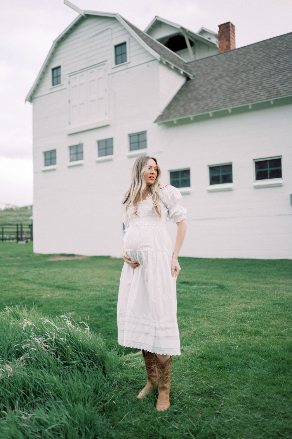 A person in a flowing white dress and brown cowboy boots stands laughing on grass near a white barn building.