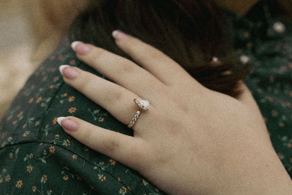 A delicate diamond engagement ring sparkles on a hand resting against a dark floral patterned dress.