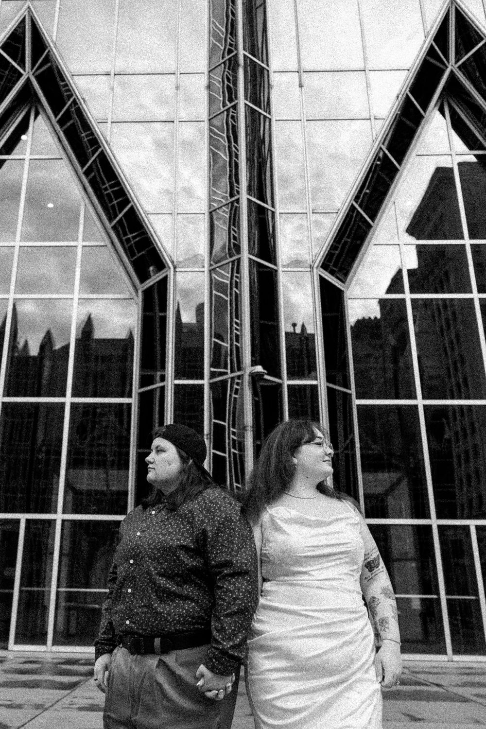 Black and white photo of two people standing back-to-back in front of a modern church building with dramatic Gothic windows.