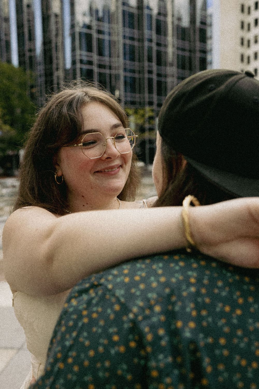Two friends share a warm embrace outdoors with city buildings visible in the background on a sunny day.
