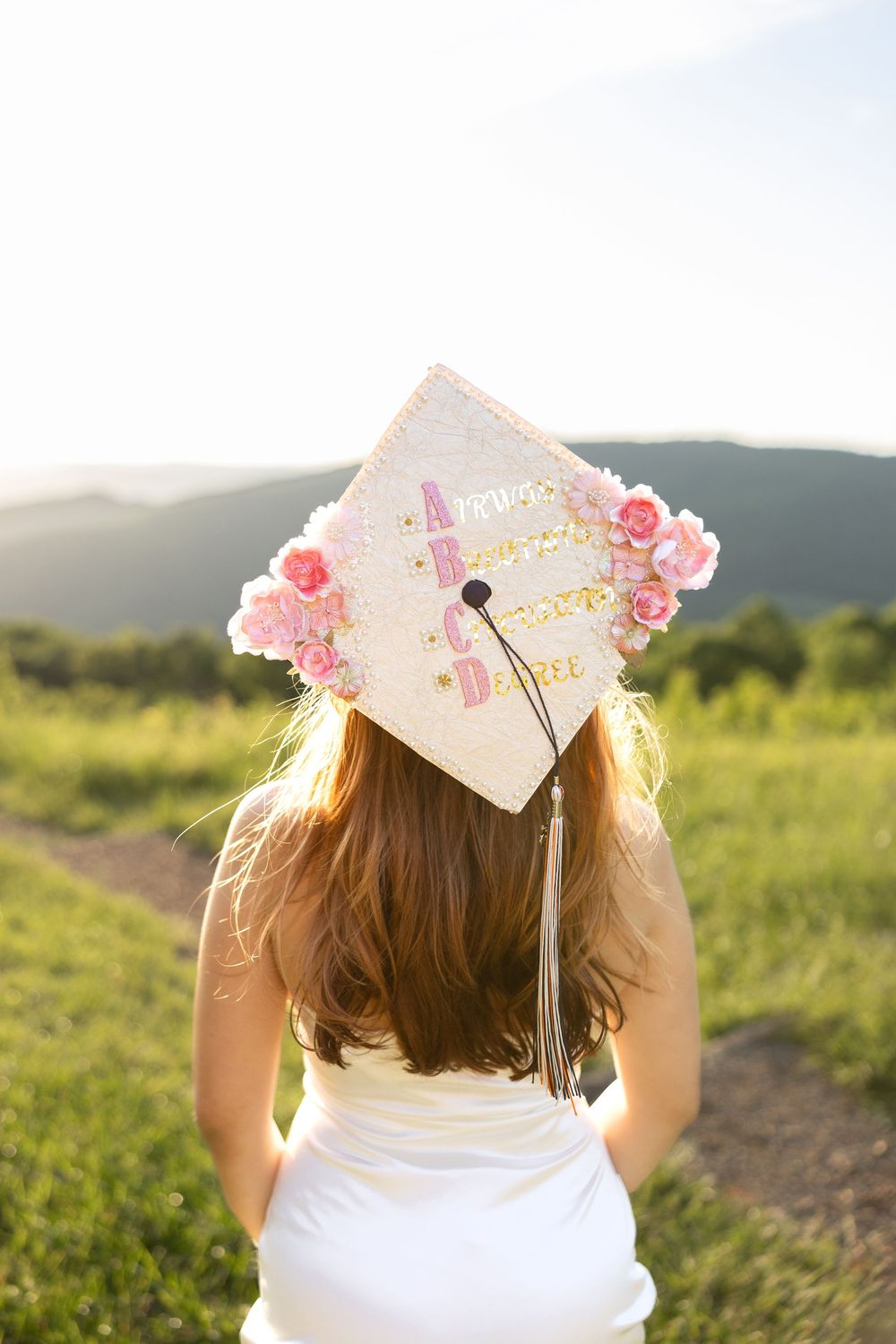 Graduate wearing white dress and decorated graduation cap with pink flowers poses outdoors against scenic mountain backdrop.