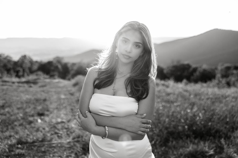 Black and white portrait of a person in a strapless dress standing in a meadow with mountains in the background during sunset.