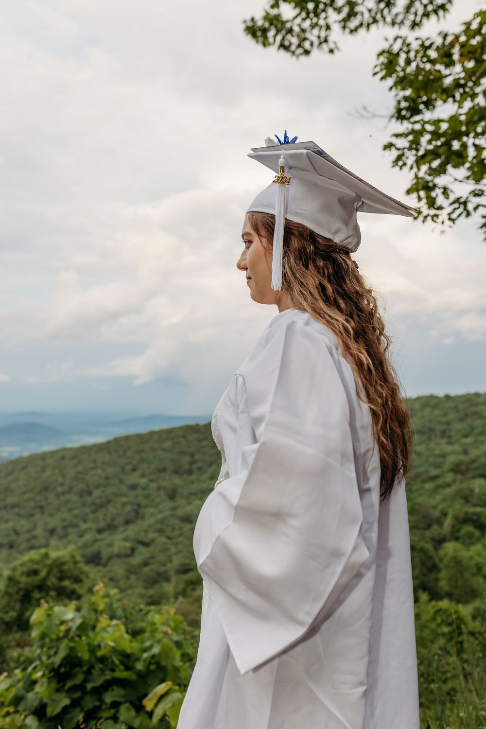 Graduate in white cap and gown stands on mountaintop overlooking scenic green rolling hills on cloudy day.