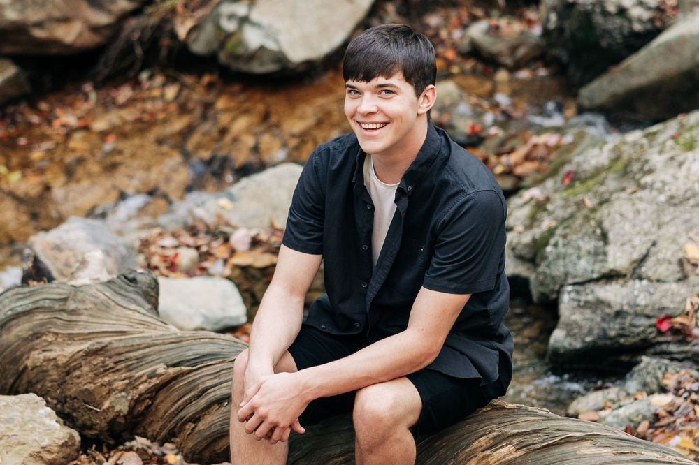 Smiling person in black shirt sitting on rocky terrain surrounded by autumn leaves.