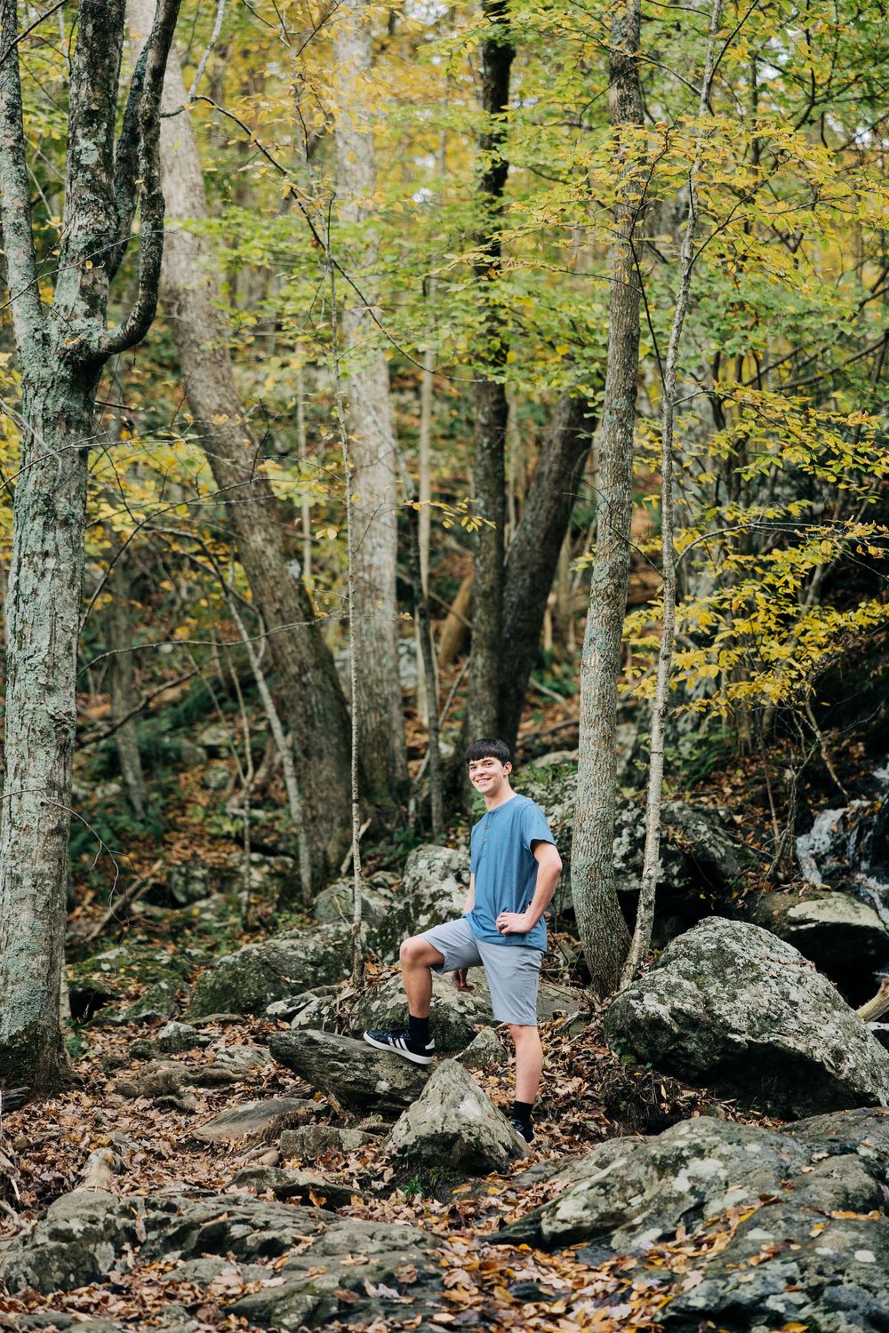 A hiker stands among large boulders and autumn trees on a rocky forest trail covered in fallen leaves.