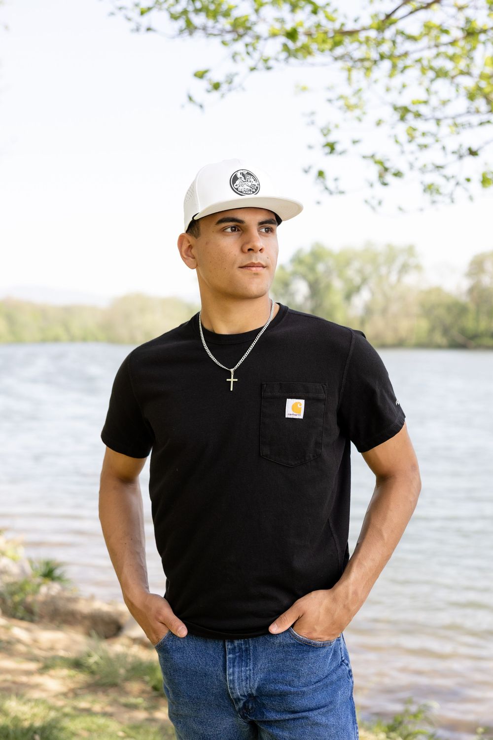 Young person in a white cap and black Carhartt t-shirt posing outdoors by a lake.