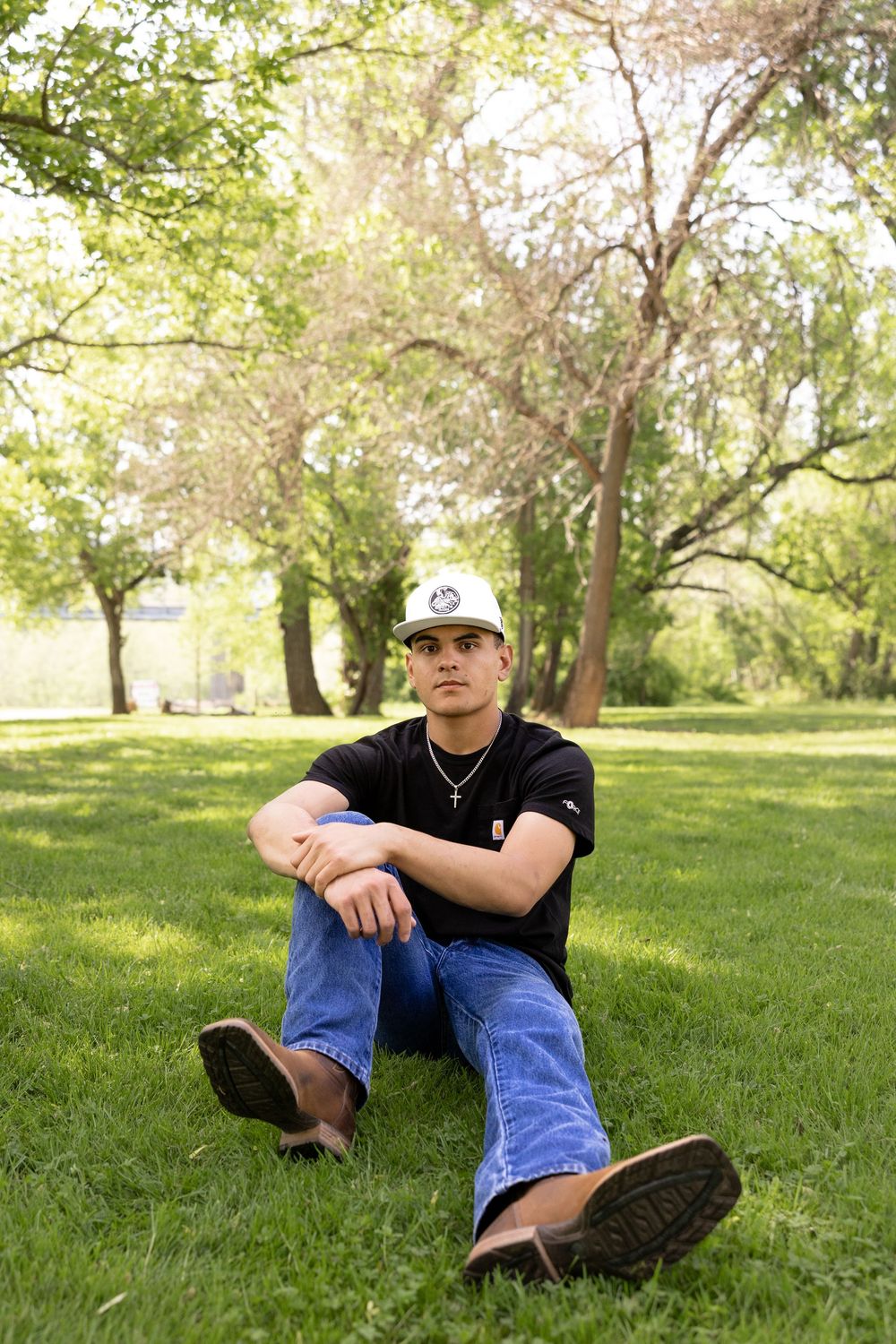 Someone wearing a white cap and black t-shirt sits relaxing on green grass in a sunny park setting.