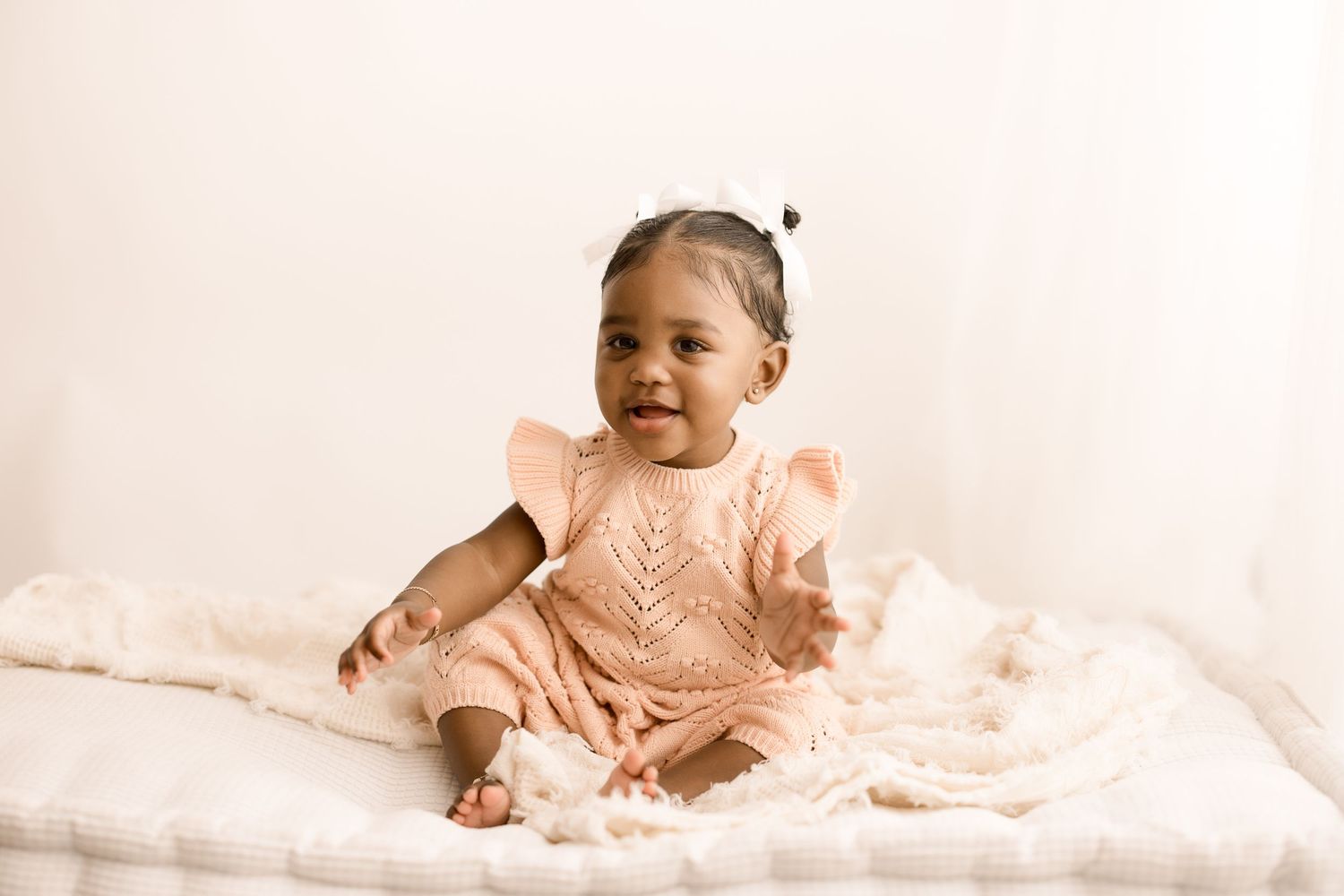 Baby wearing peach-colored ruffled dress sits on white textured bedding in warm neutral studio setting.