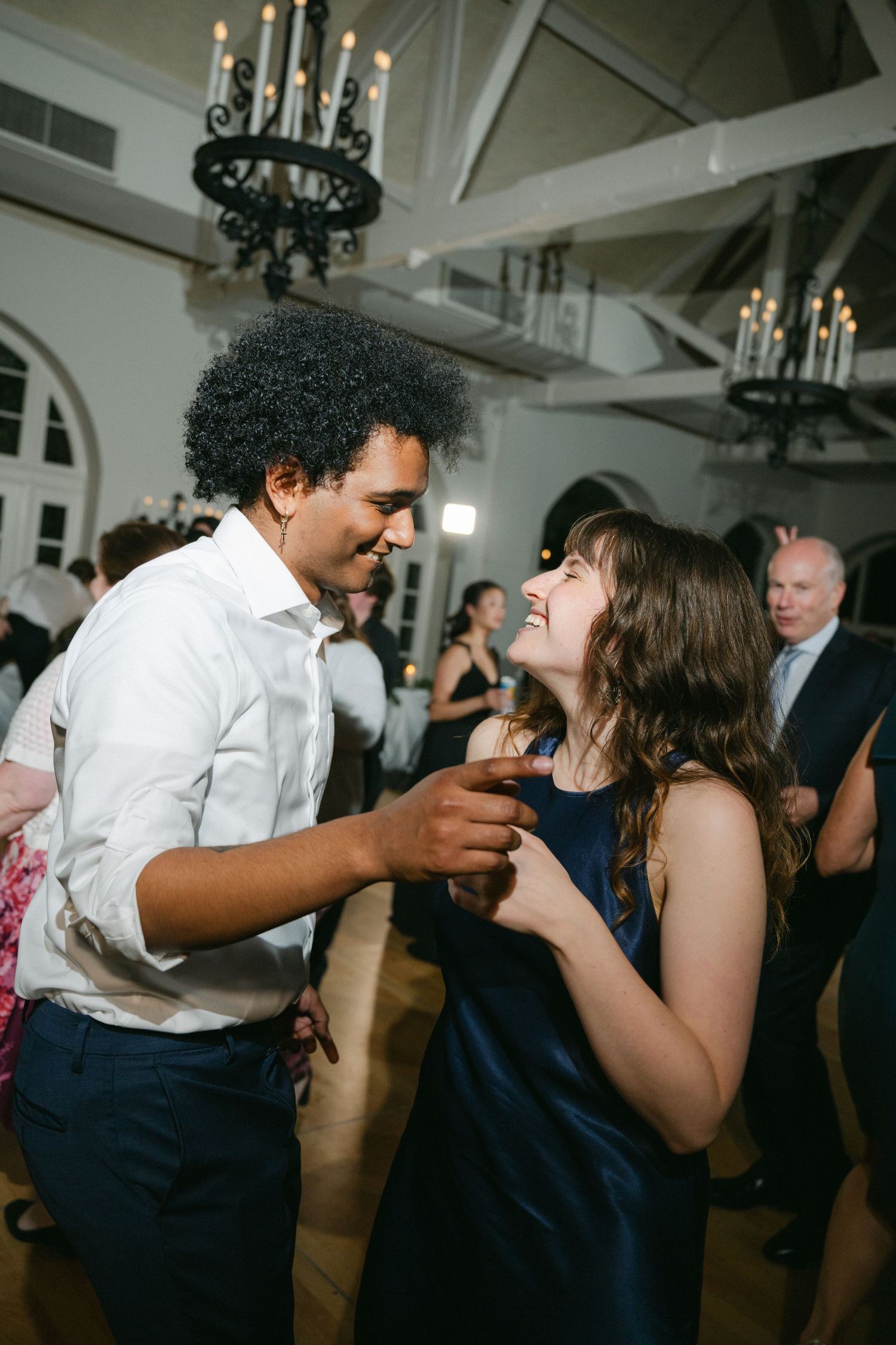 Two wedding guests share an energetic moment dancing together at a reception under elegant chandeliers.