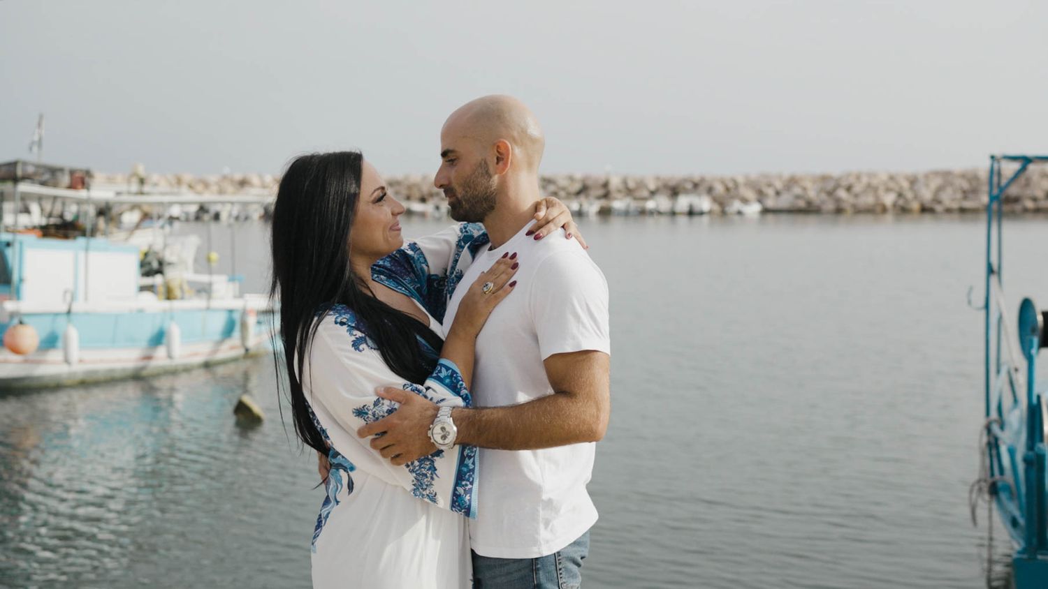 Romantic couple embraces on a dock by the marina with boats and calm water in the background.