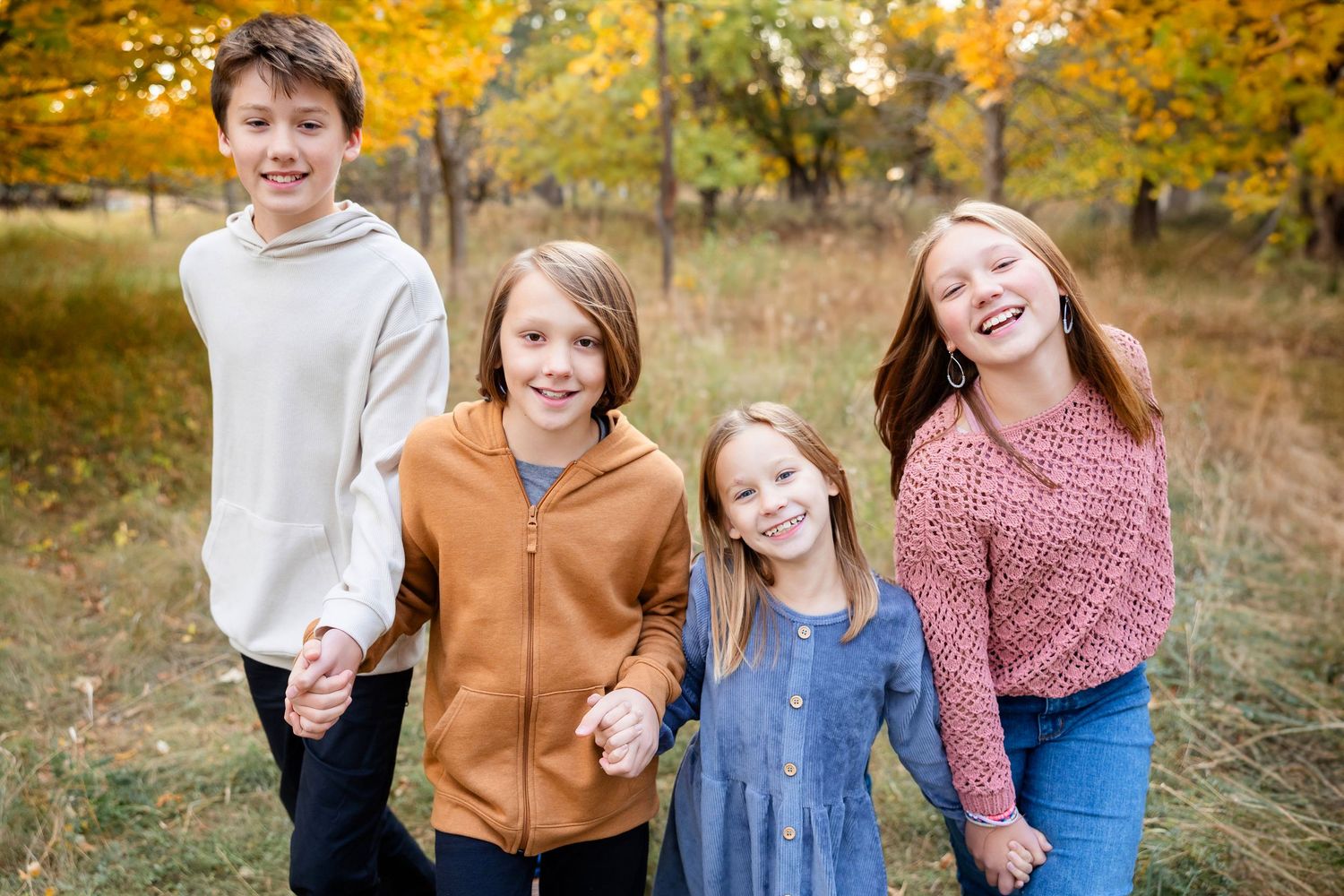 Four children stand together smiling outdoors with autumn foliage and golden leaves in the background.