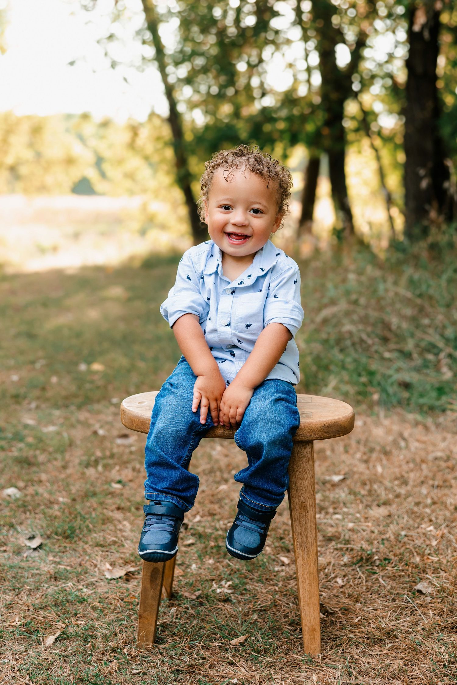 A small child wearing a light blue button-down shirt and jeans sits on a wooden stool outdoors in dappled sunlight.